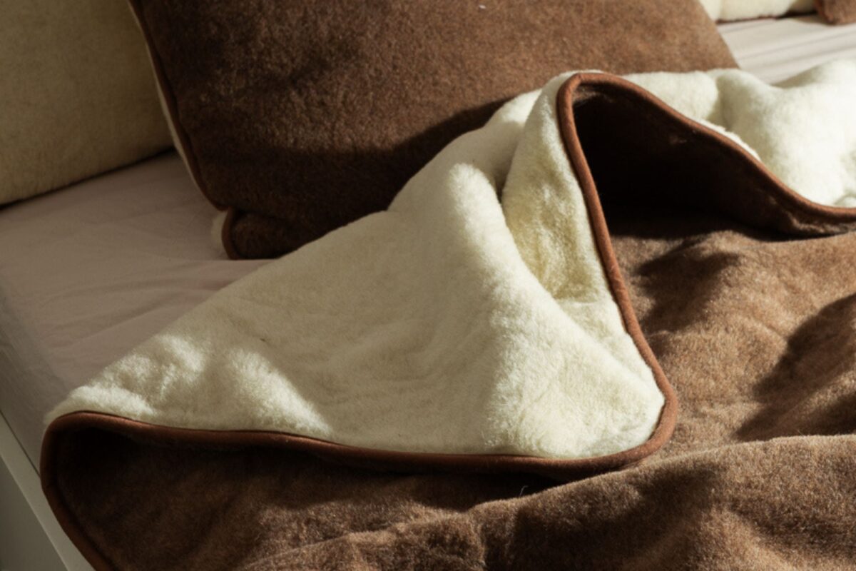 Zbliżenie na łóżko na którym leży odkryta kołdra wełniana dwustronna - z jednej strony brązowa, z drugiej ecru. W tle poduszki z wełny  brązowe i ecuru.